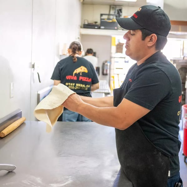 A Viva Pizza employee in a black cap and apron stretches pizza dough in a commercial kitchen.