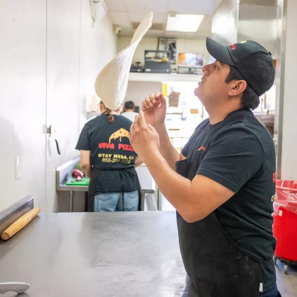 A Viva Pizza chef in a black cap and apron expertly tosses pizza dough high in the air.