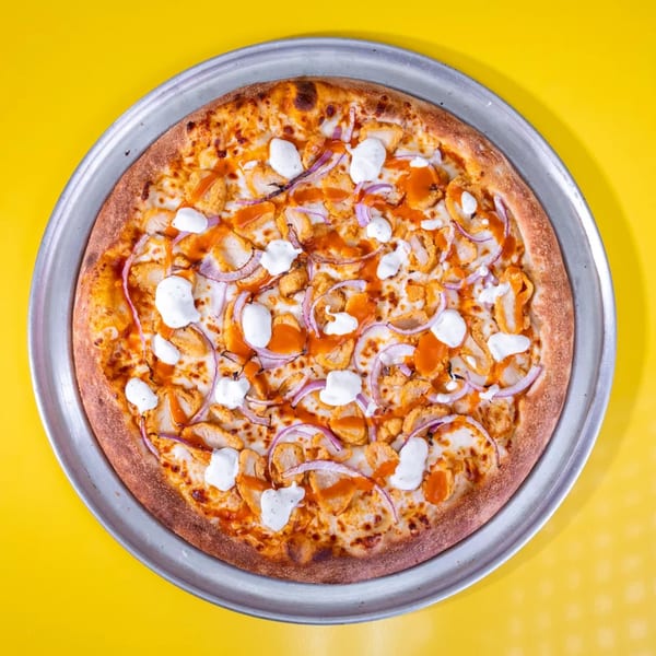 Buffalo chicken pizza with red onions and ranch drizzle on a metal pan, against a yellow background.