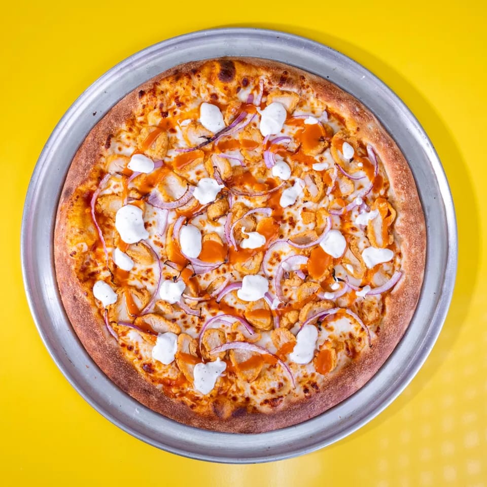 Buffalo chicken pizza with red onions and ranch drizzle on a metal pan, against a yellow background.