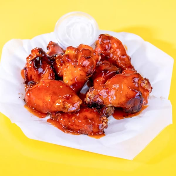 A basket of glossy, red-sauced chicken wings with a side of ranch dressing on a yellow background.