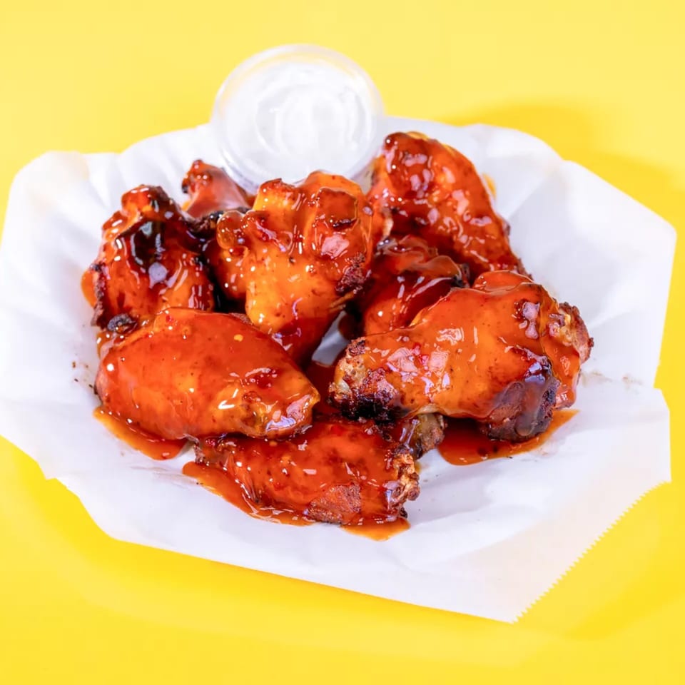 A basket of glossy, red-sauced chicken wings with a side of ranch dressing on a yellow background.
