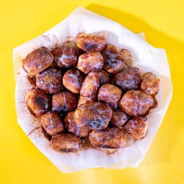 A pile of cinnamon sugar donut holes with white glaze in a paper-lined basket on a yellow background.